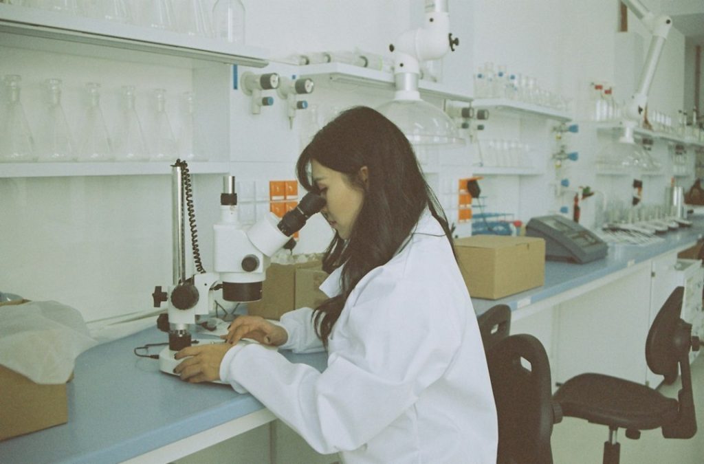 Farmacêutica de jaleco branco trabalhando ao microscópio em laboratório limpo