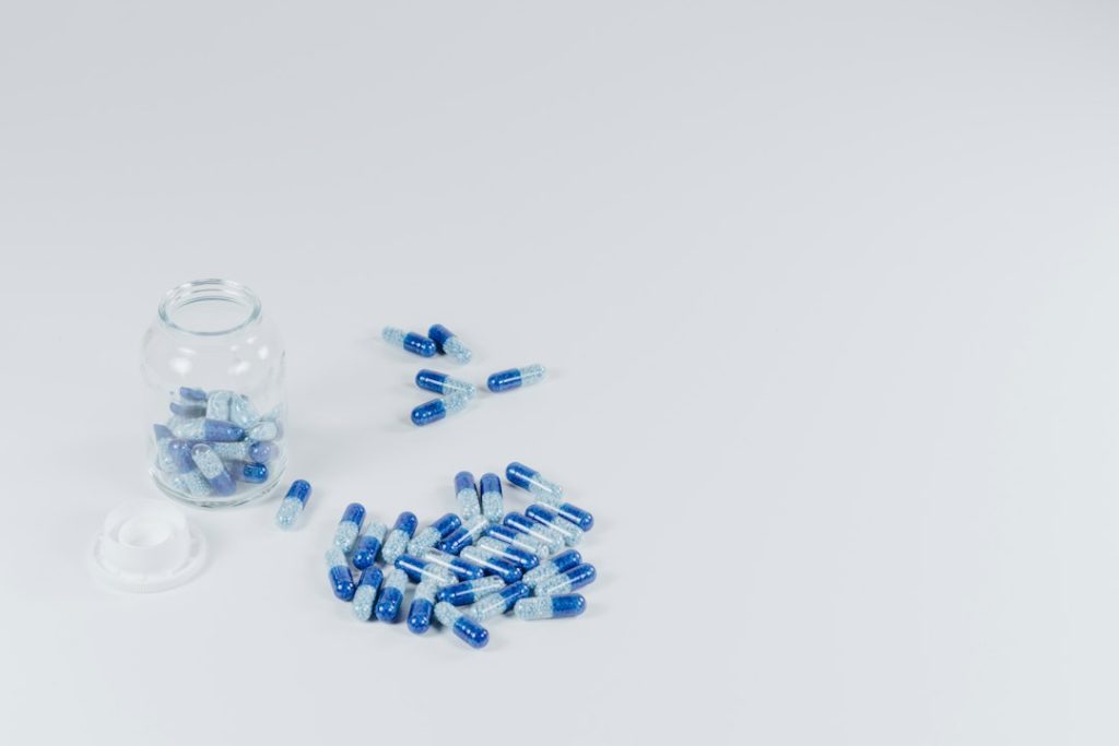 Frasco de vidro com tampa aberta e várias cápsulas azuis e brancas espalhadas sobre superfície branca.