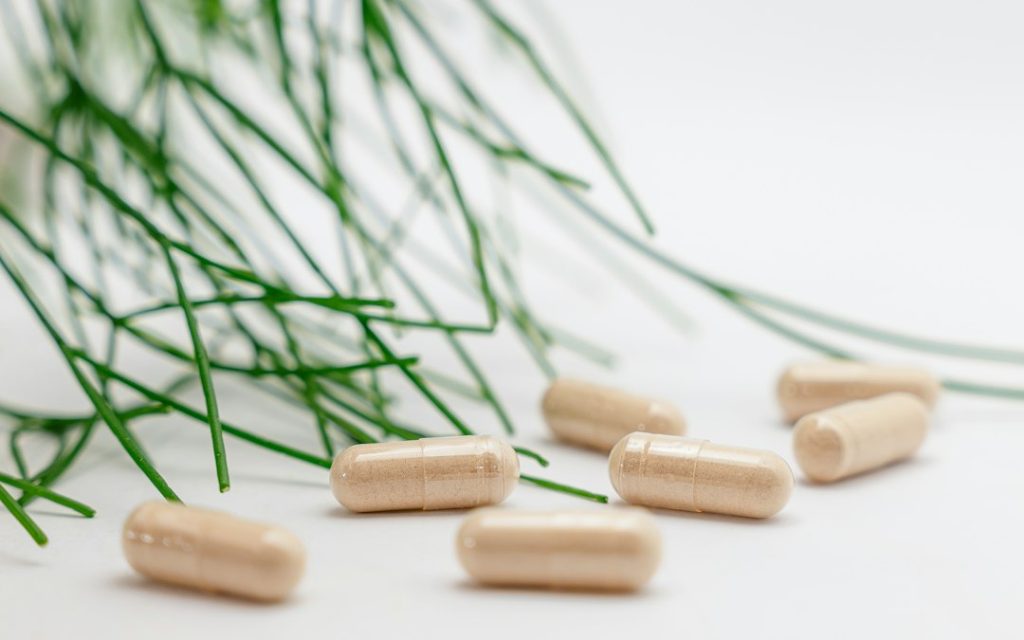 Cápsulas de suplementos em cima de uma superfície branca com ramos verdes ao fundo.