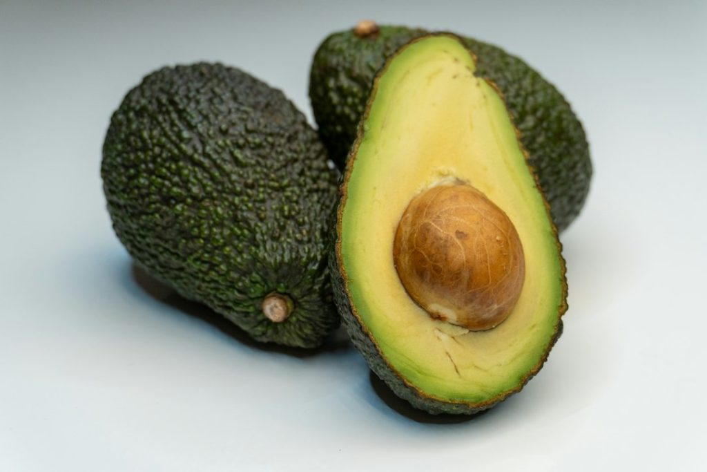 Metade de um abacate fresco com caroço e dois abacates inteiros sobre fundo claro.