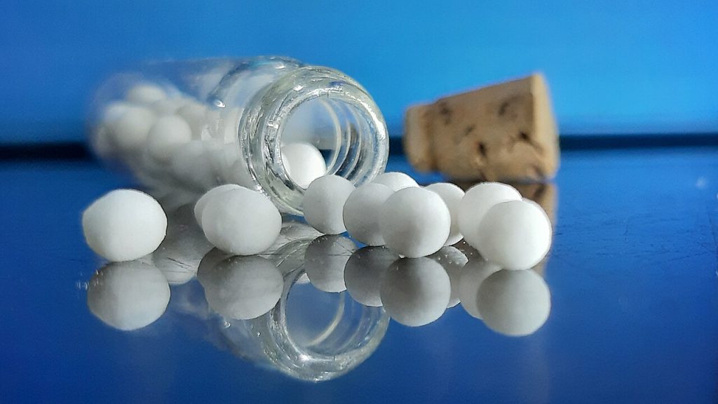 Glóbulos homeopáticos brancos saindo de um frasco de vidro transparente sobre superfície reflexiva azul
