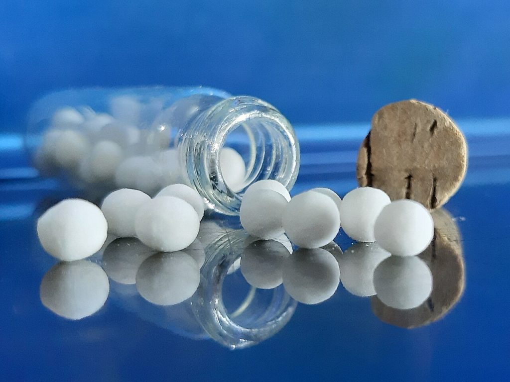 Pequenas pílulas homeopáticas brancas saindo de um frasco de vidro com rolha sobre uma superfície reflexiva azul.