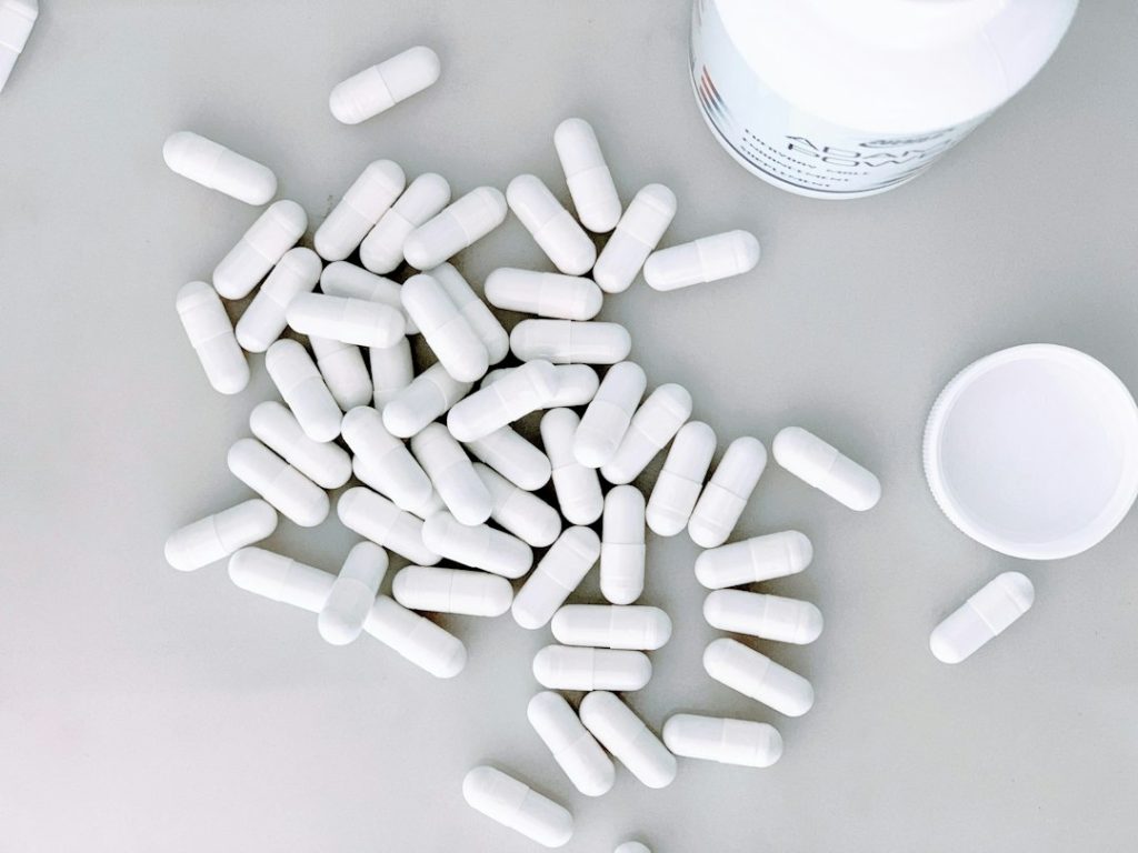 Cápsulas de suplementos manipulados espalhadas ao lado de um frasco branco em fundo neutro.