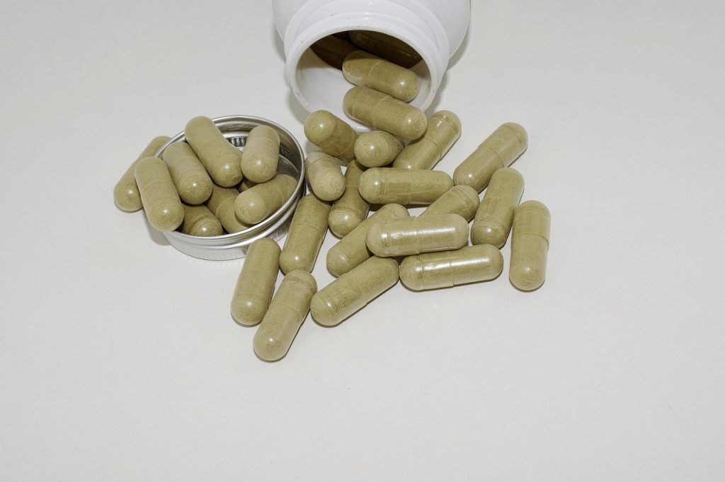 Cápsulas de suplemento verde-oliva derramadas de um frasco branco sobre bancada.