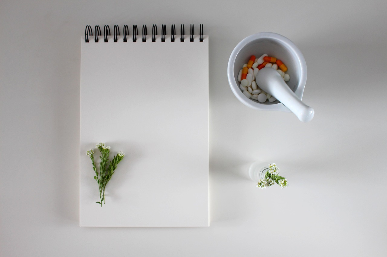 Caderno de anotações em cima de mesa branca ao lado de pilão com cápsulas e comprimidos e flores brancas.