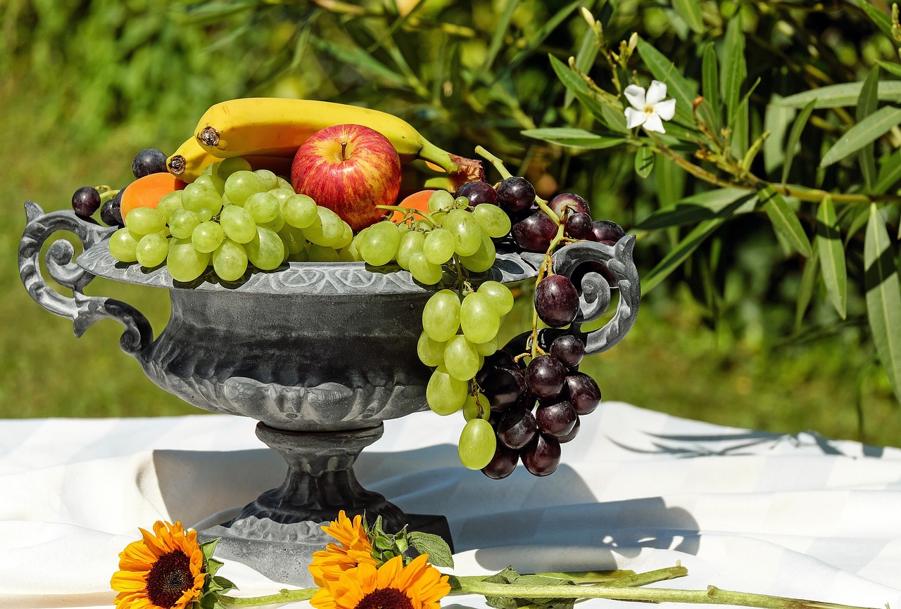 Fruteira com frutas frescas como bananas, maçãs e uvas sobre uma mesa branca ao ar livre