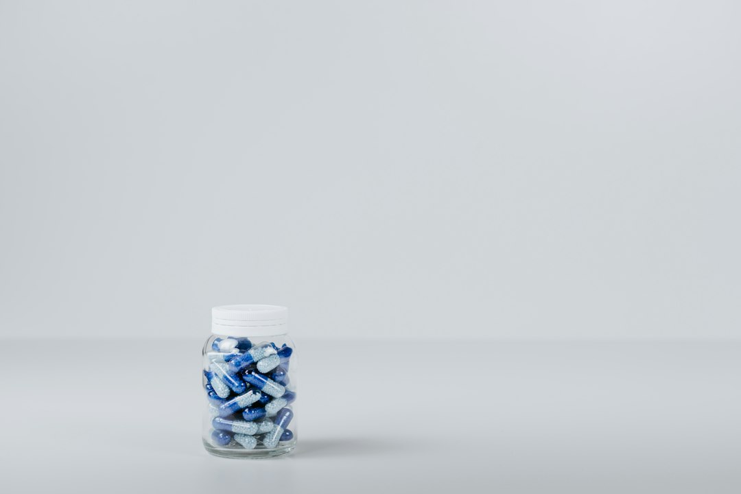 Frasco transparente com tampa branca cheio de cápsulas azul e branco sobre bancada clara.