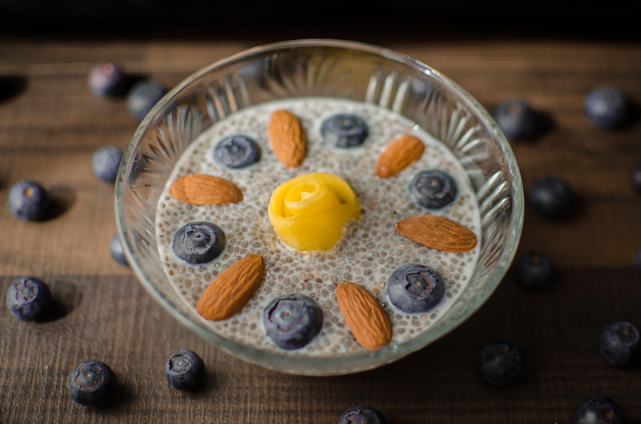 Bowl de vidro com pudim de chia, leite, mirtilos e amêndoas ao redor de uma flor de manga em mesa de madeira.