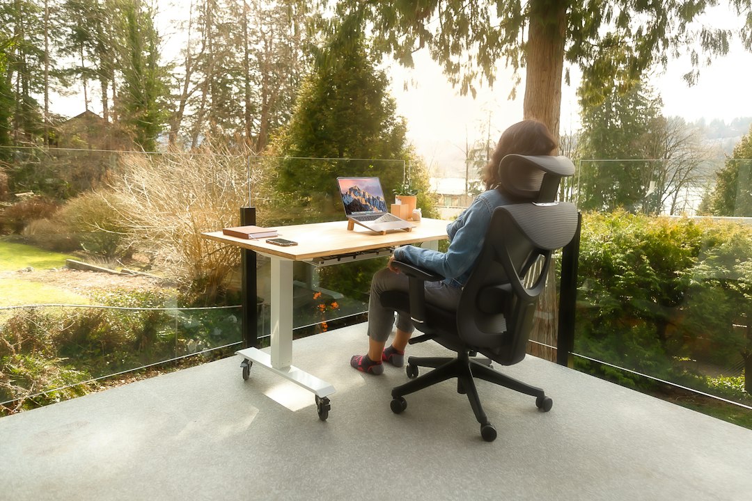 Mulher trabalhando em home office com cadeira ergonômica e mesa ajustável em varanda arborizada.