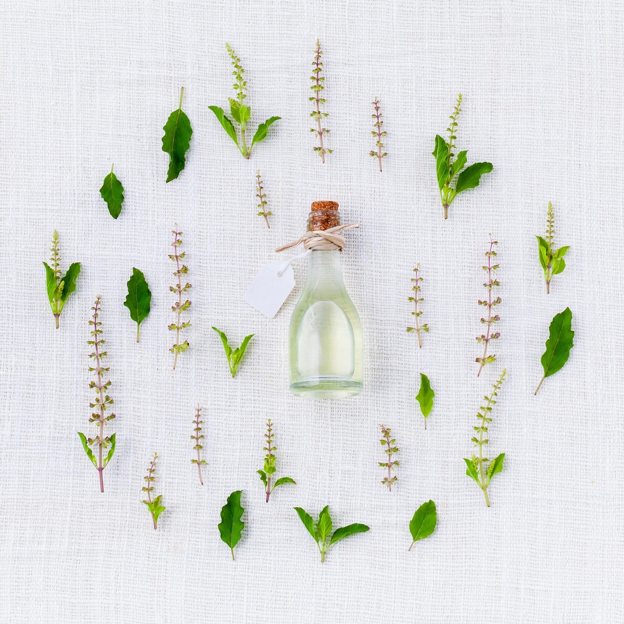 Frasco de vidro com conta-gotas cercado por folhas e flores em arranjo simétrico sobre tecido branco.