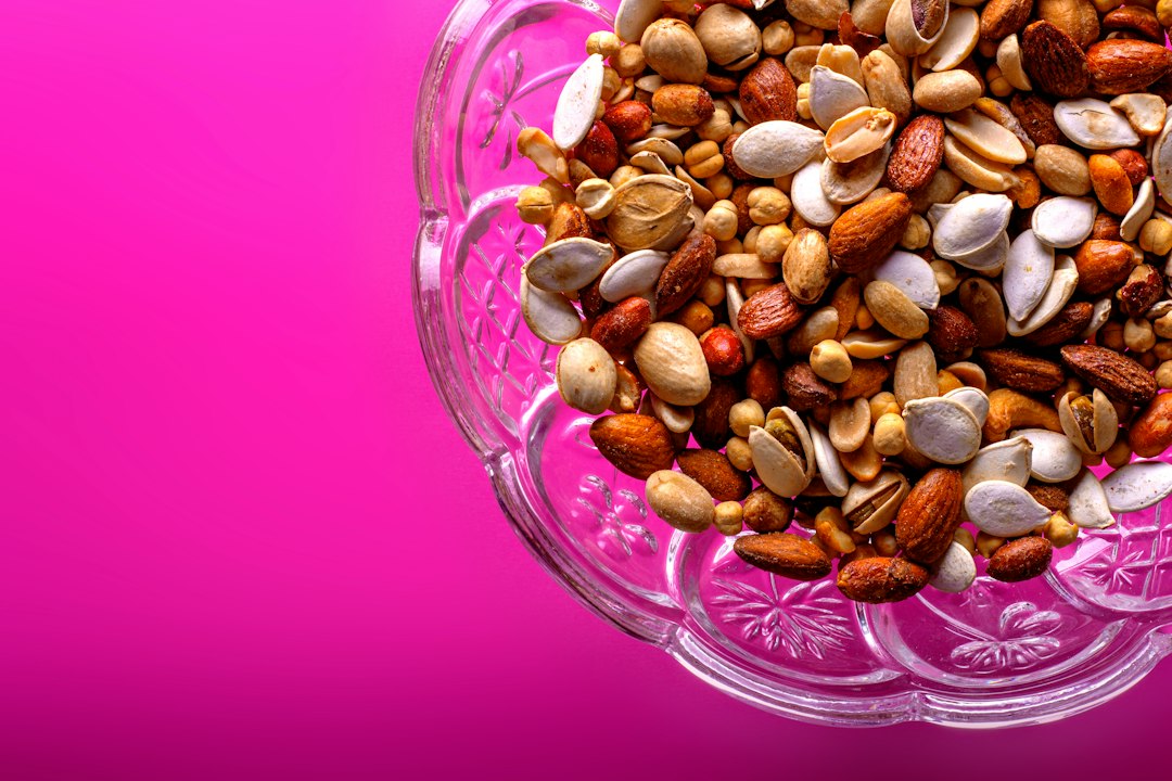 Tigela de vidro com mix de castanhas, amêndoas e sementes sobre fundo rosa.