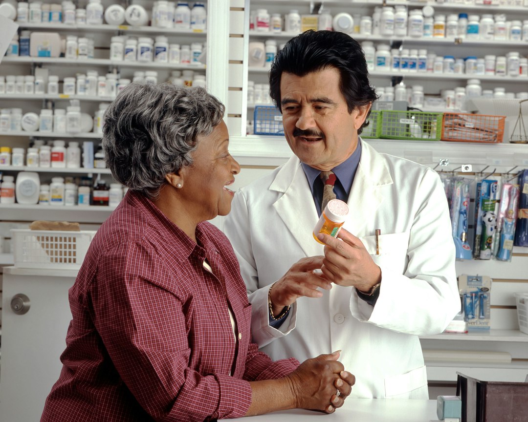 Farmacêutico conversando com uma cliente idosa e mostrando um frasco de medicamento em uma farmácia.